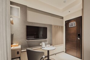 Habitación de hotel moderna con televisor de pantalla plana, una pequeña mesa redonda con tetera y tazas, una silla y un armario con un cartel de número de puerta.