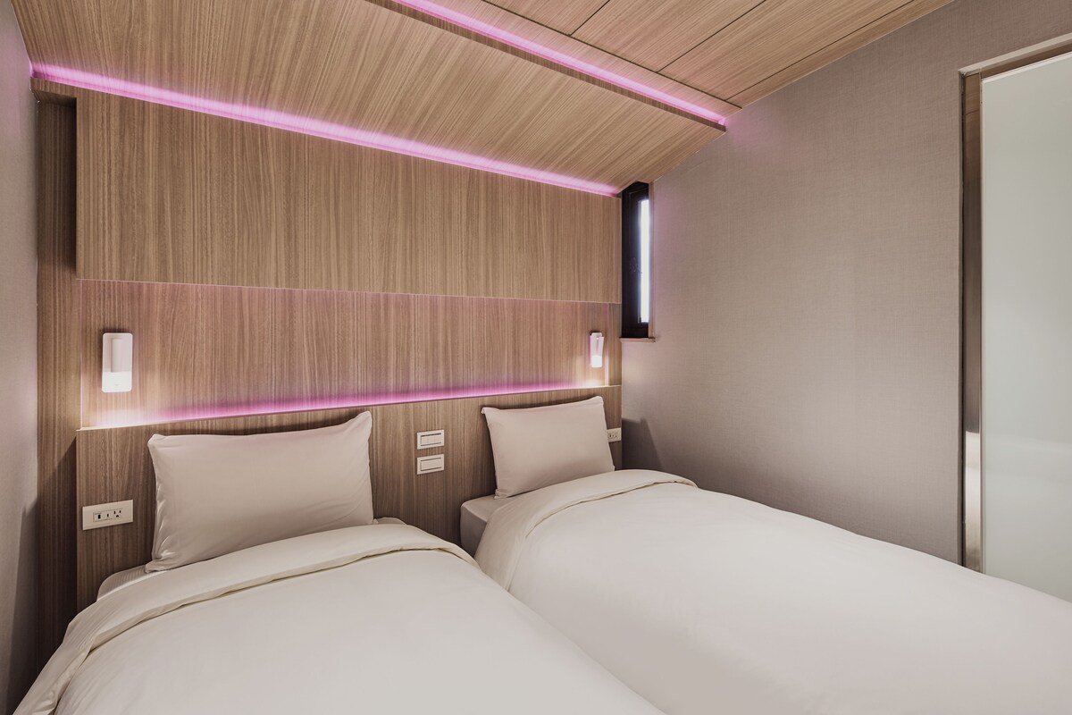 A modern hotel room with two beds, wooden paneling, and built-in lighting.