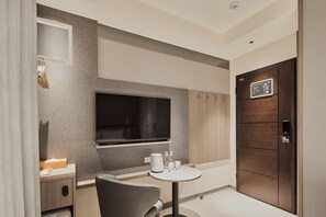 Una habitación de hotel moderna con un televisor de pantalla plana montado en la pared, una pequeña mesa redonda con una tetera y vasos, una silla gris y una puerta de madera con un cartel.