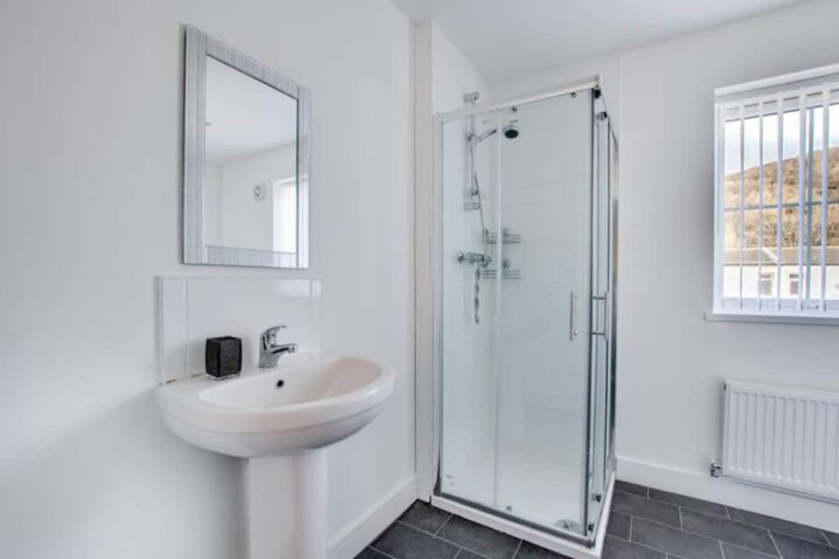 A bathroom with a white sink, a glass shower enclosure, a mirror, and a window with a view.