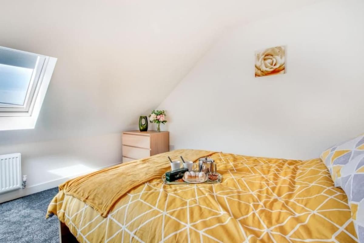 Quarto com cama, janela, radiador, mesa de cabeceira com um vaso e uma decoração na parede.