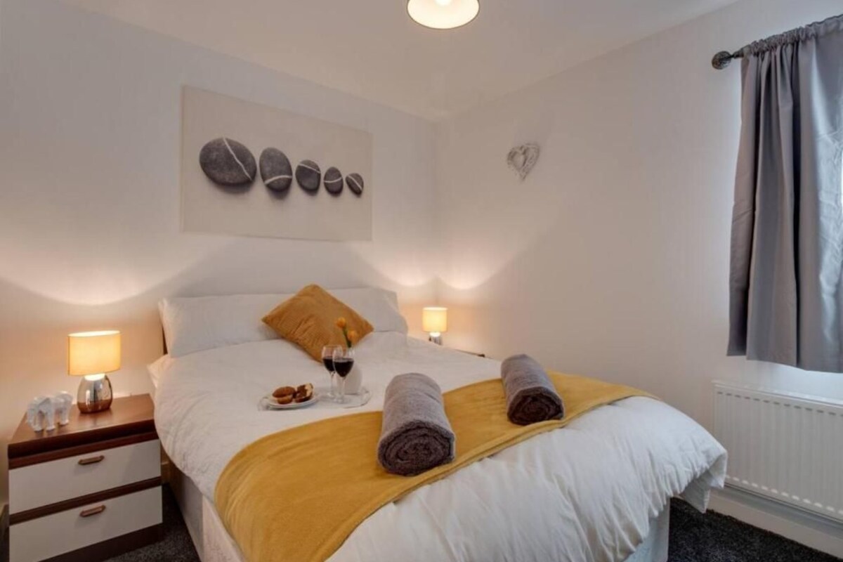 A neatly made bed with a white comforter and a yellow throw blanket, two rolled towels, a wine glass, and a plate of food on the nightstand.