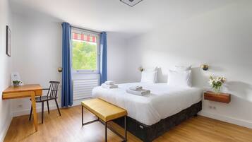 Un dormitorio con cama, escritorio, silla, banco, ventana con cortinas y mesita de noche con un jarrón.