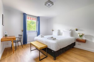 Quarto com cama, escrivaninha, cadeira, banco, janela com cortinas e mesa de cabeceira com um vaso.