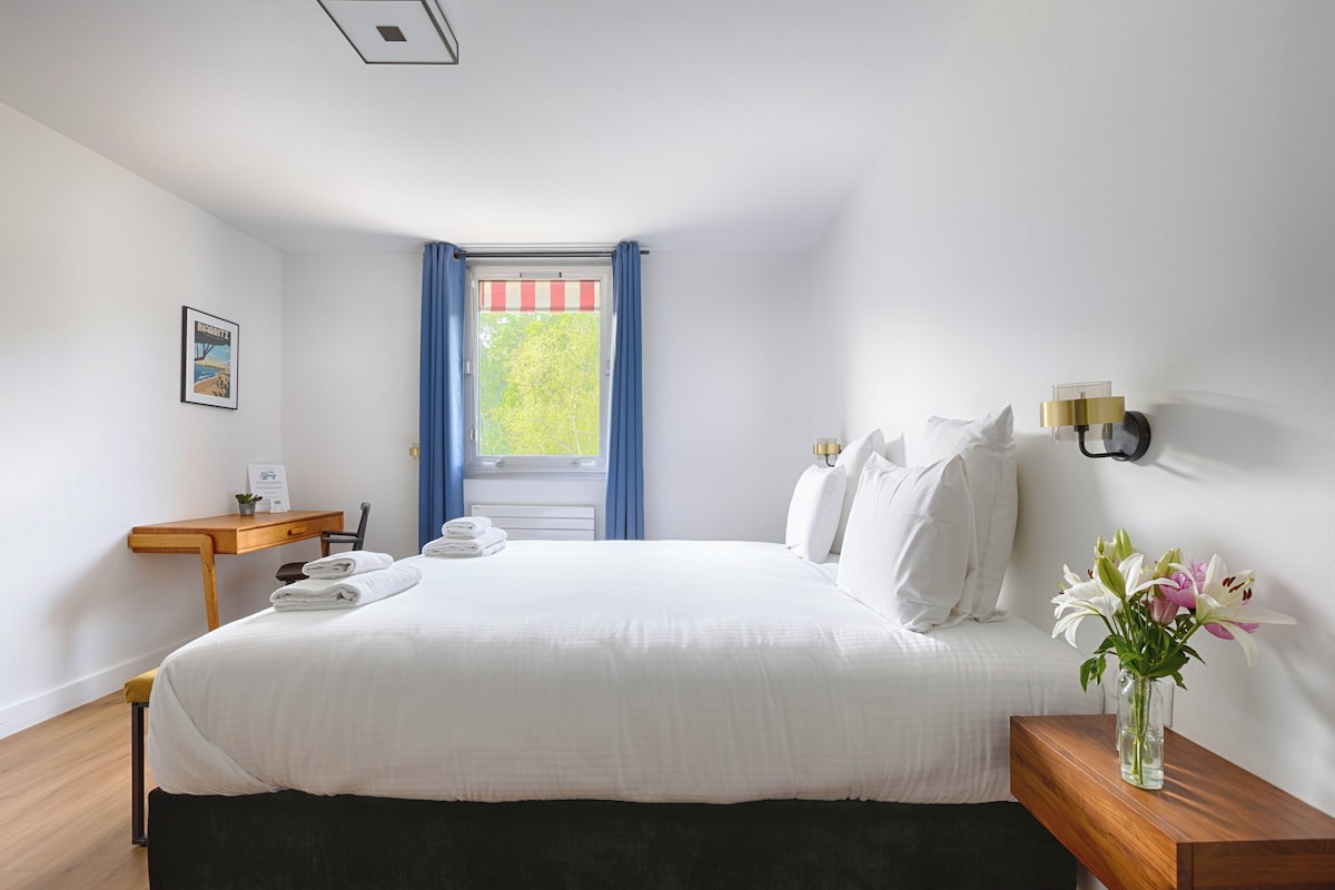 A hotel room with a bed, a desk, a chair, a nightstand, a vase with flowers, and a window with curtains.