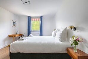 Habitación de hotel con cama, escritorio, silla, mesita de noche, un jarrón con flores y una ventana con cortinas.
