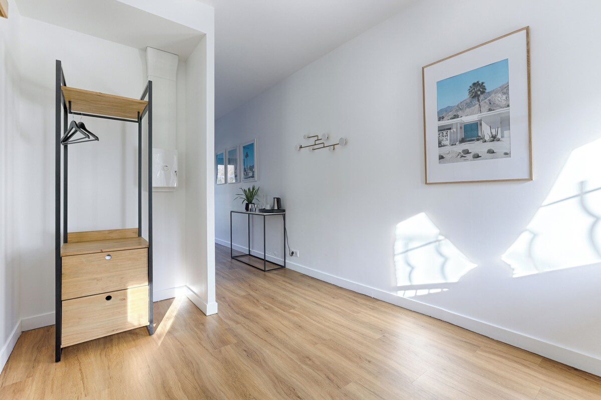 A room with a wooden floor, a metal-framed wardrobe, a small table with a plant, and a framed picture on the wall.