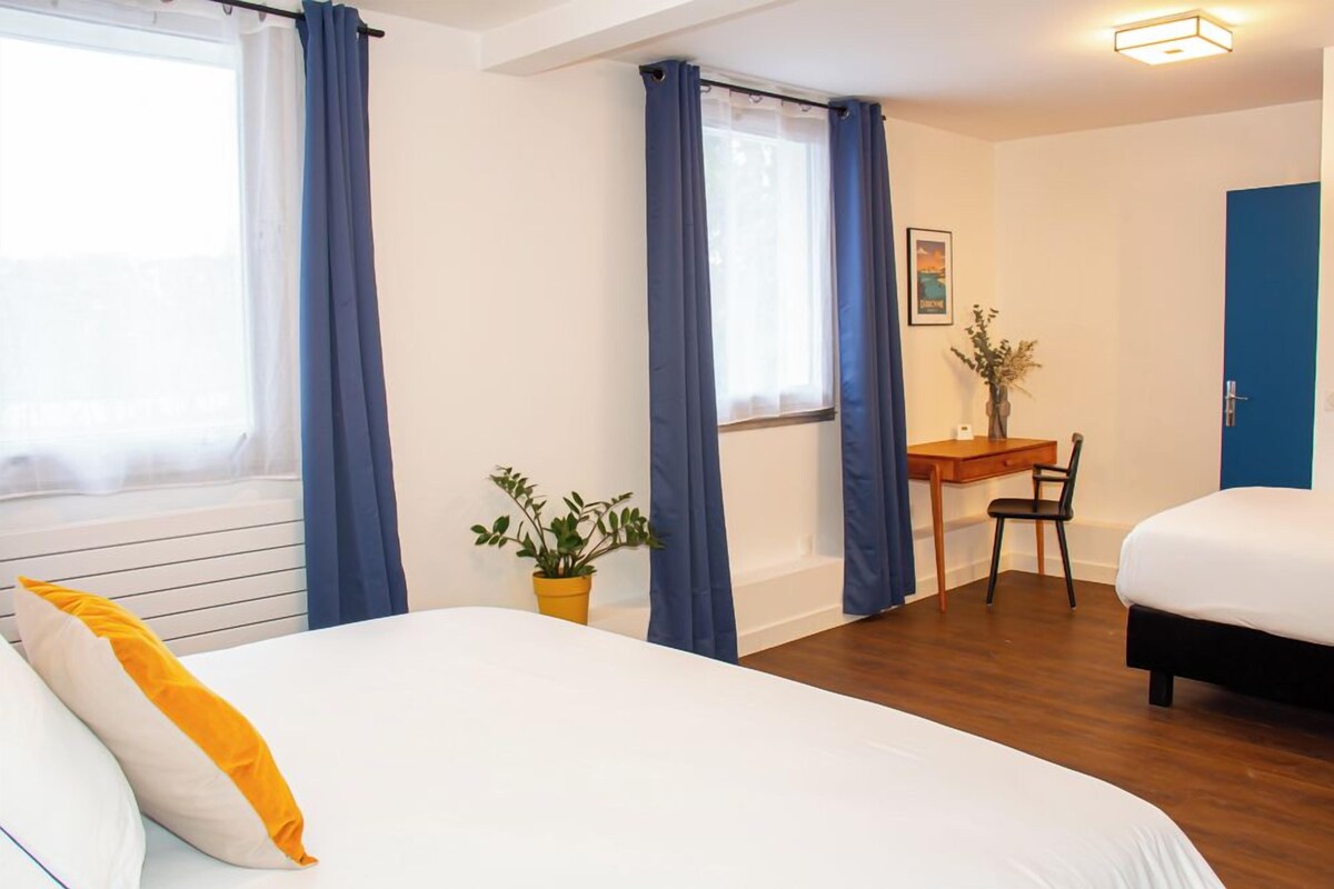 A hotel room with a bed, a desk, a chair, a potted plant, and a door.