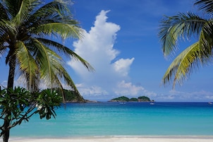 Una playa con agua turquesa cristalina, palmeras y una isla distante.