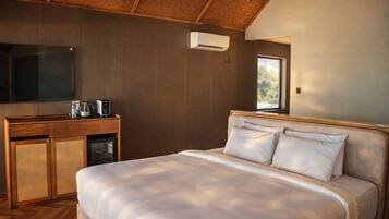 A bedroom with a bed, wooden headboard, a TV mounted on the wall, a small kitchenette with a microwave, and a window providing natural light.