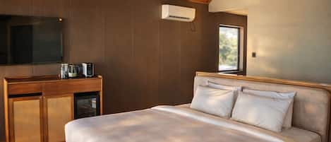 Un dormitorio con cama, cabecera de madera, televisor montado en la pared, una pequeña kitchenette con microondas y una ventana que proporciona luz natural.