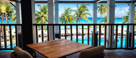 A wooden table with two chairs, a view of a poolside area with palm trees and a beach, and a clear sky.