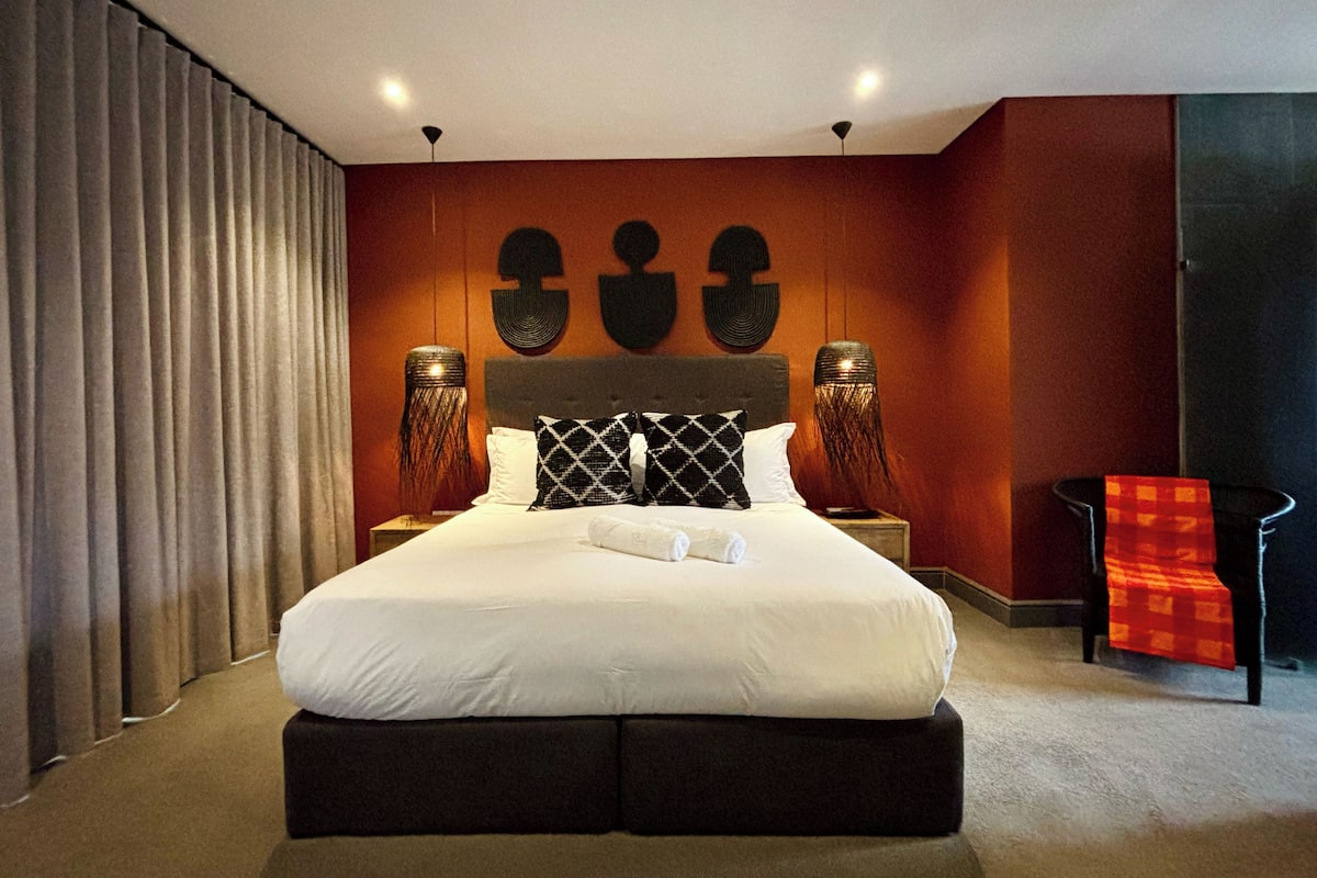 A modern hotel room with a large bed, dark headboard, two bedside tables, and a red chair.