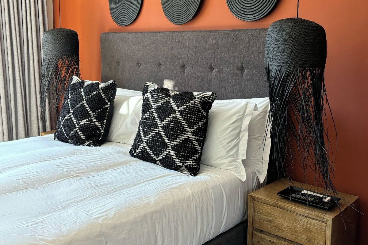 A bed with white linens, two patterned pillows, a grey headboard, and a wooden nightstand with a remote control.