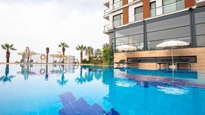 A swimming pool with lounge chairs and umbrellas, a modern building with balconies, and palm trees. - Maia Beach Hotel (Aydın)