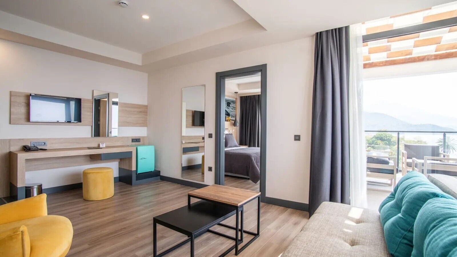 A modern hotel room with a bed, a desk, a TV, a small kitchenette, and a balcony with a view.