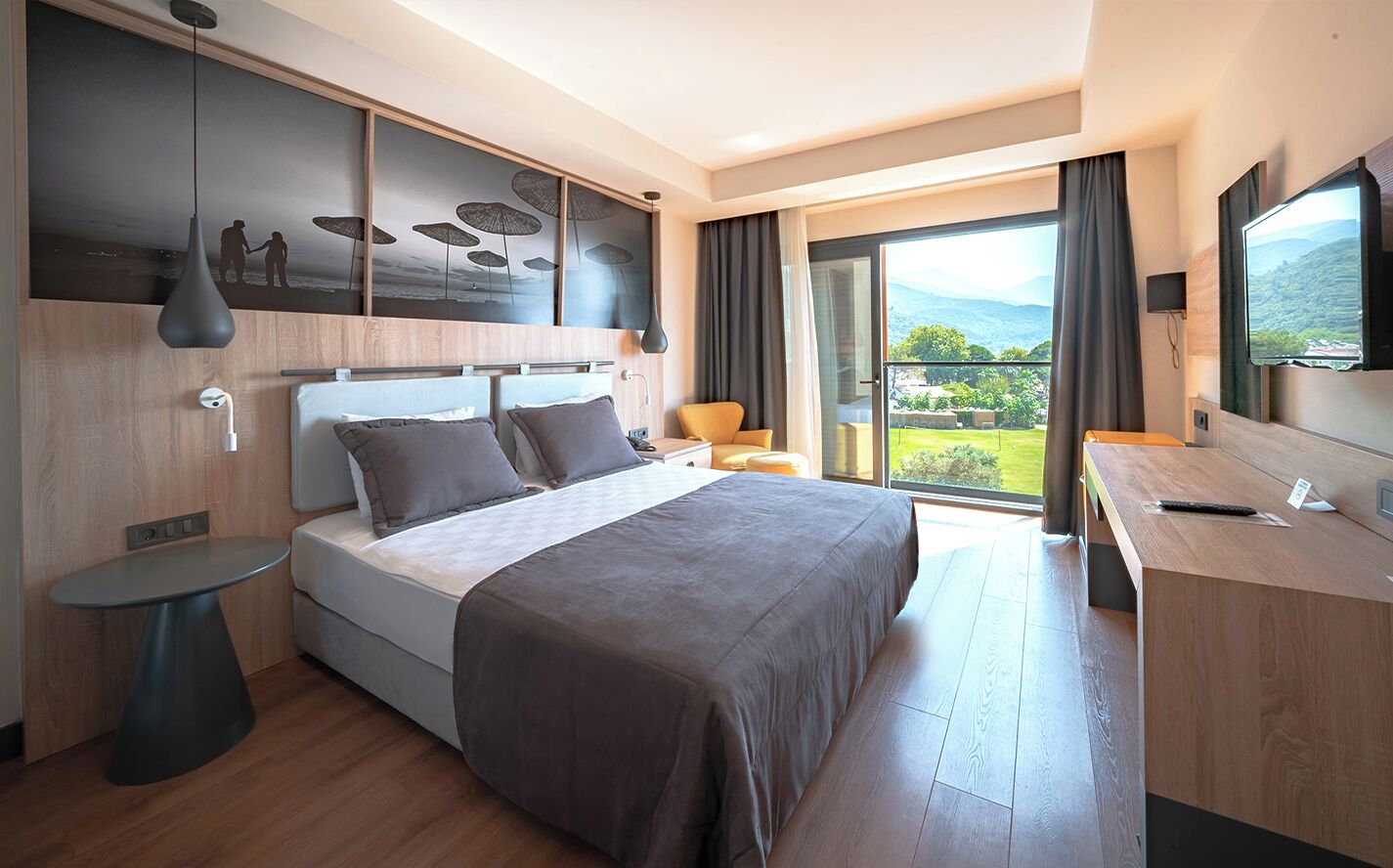 A modern hotel room with a large bed, a desk, a chair, a TV, and a balcony with a mountain view.