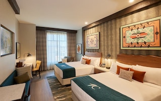 Una habitación de hotel con dos camas, un escritorio, una silla y un ventanal con cortinas.