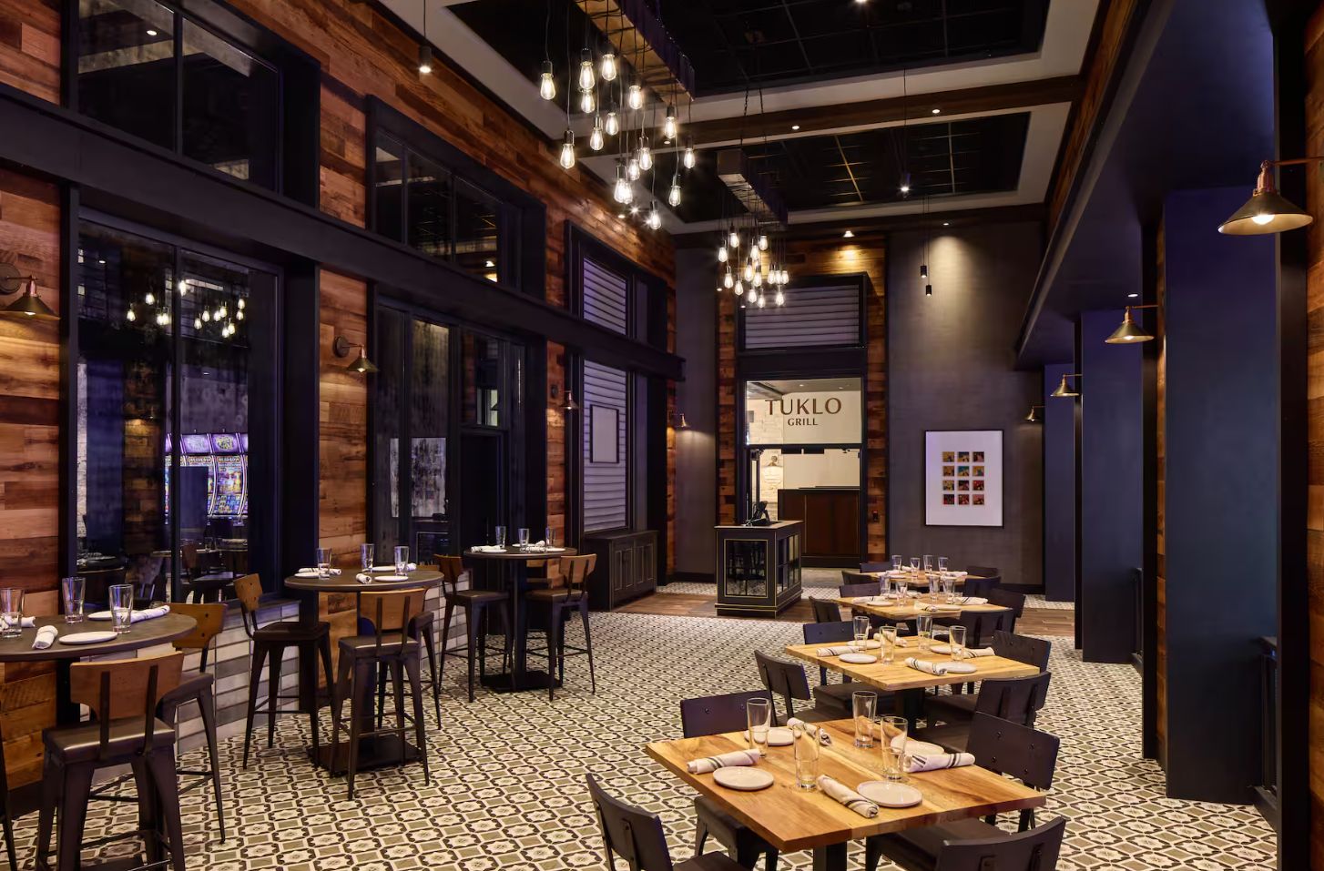 A restaurant interior with wooden walls, hanging light fixtures, and a patterned floor. Tables are set with plates and glasses.