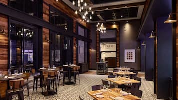 A restaurant interior with wooden walls, hanging light fixtures, and a patterned floor. Tables are set with plates and glasses.