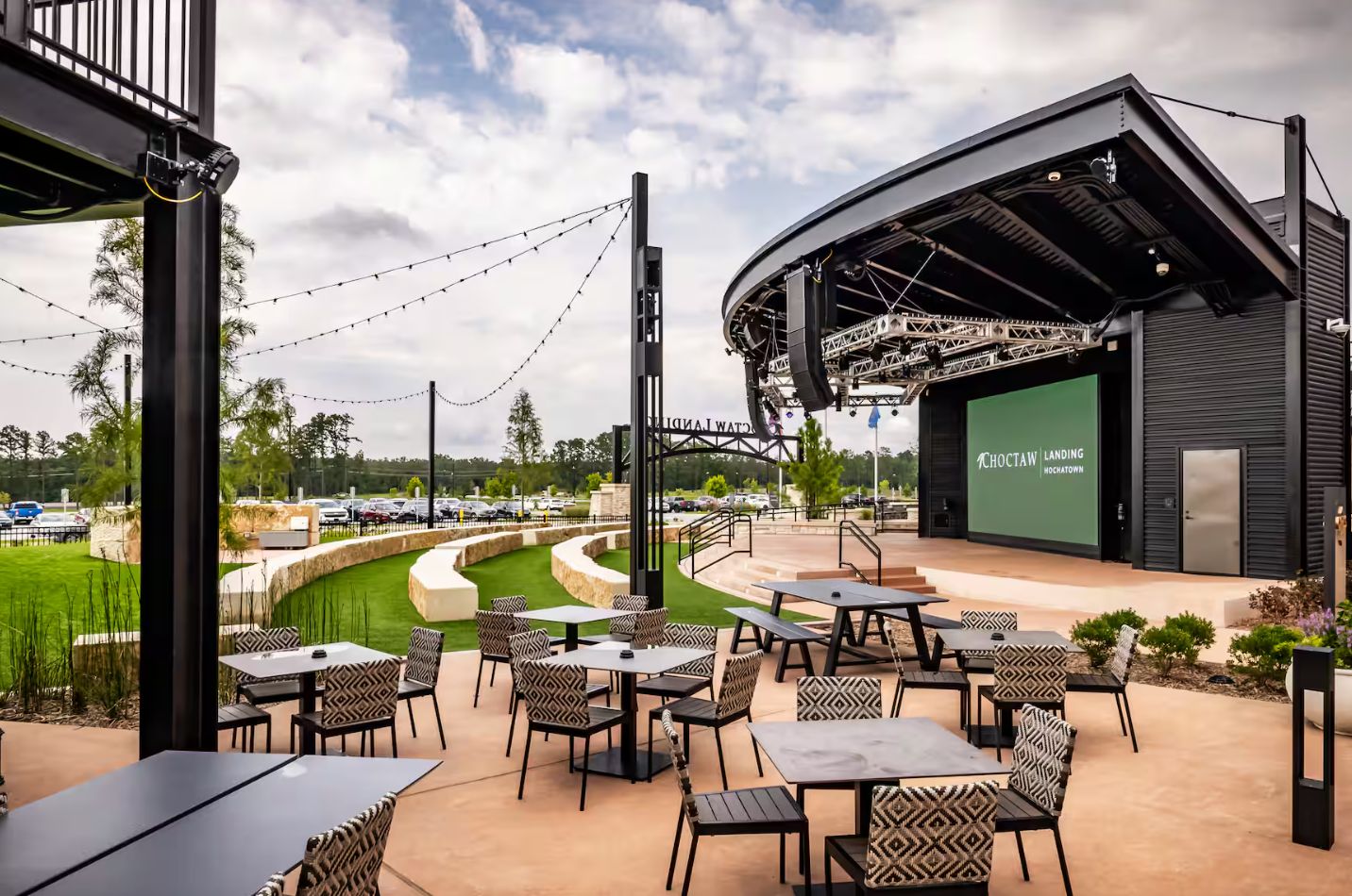 An outdoor patio area with tables and chairs, a stage with a screen, and a view of a parking lot.