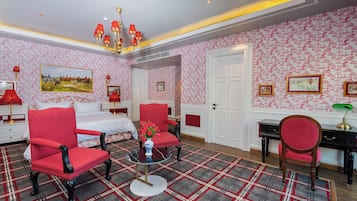 A hotel room with a bed, two red armchairs, a small table with a vase, a desk with a lamp, and a patterned carpet.
