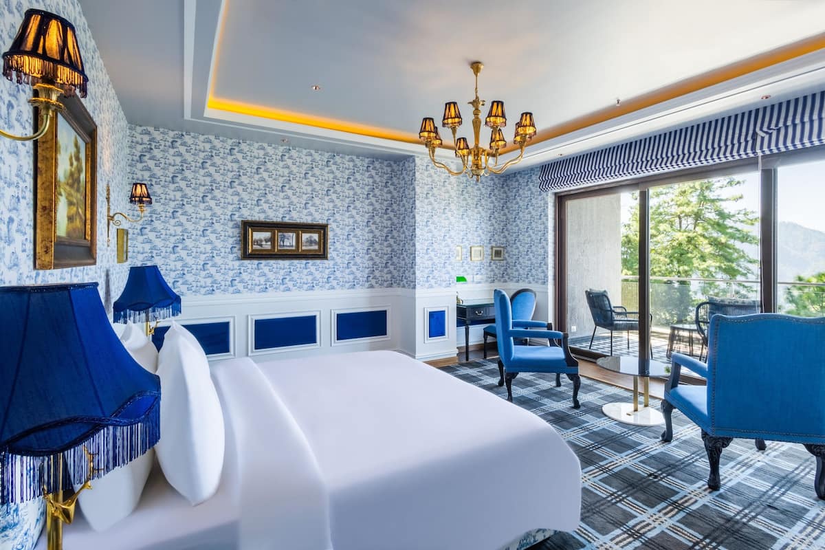 A hotel room with a bed, blue chairs, a desk, and a view of the outdoors.