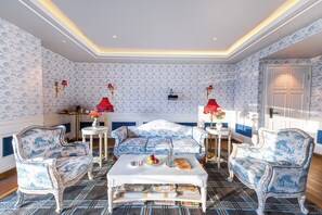 Una habitación con papel tapiz estampado, un sofá blanco, dos sillones azules, una mesa de centro con frutas y un jarrón, y lámparas rojas.