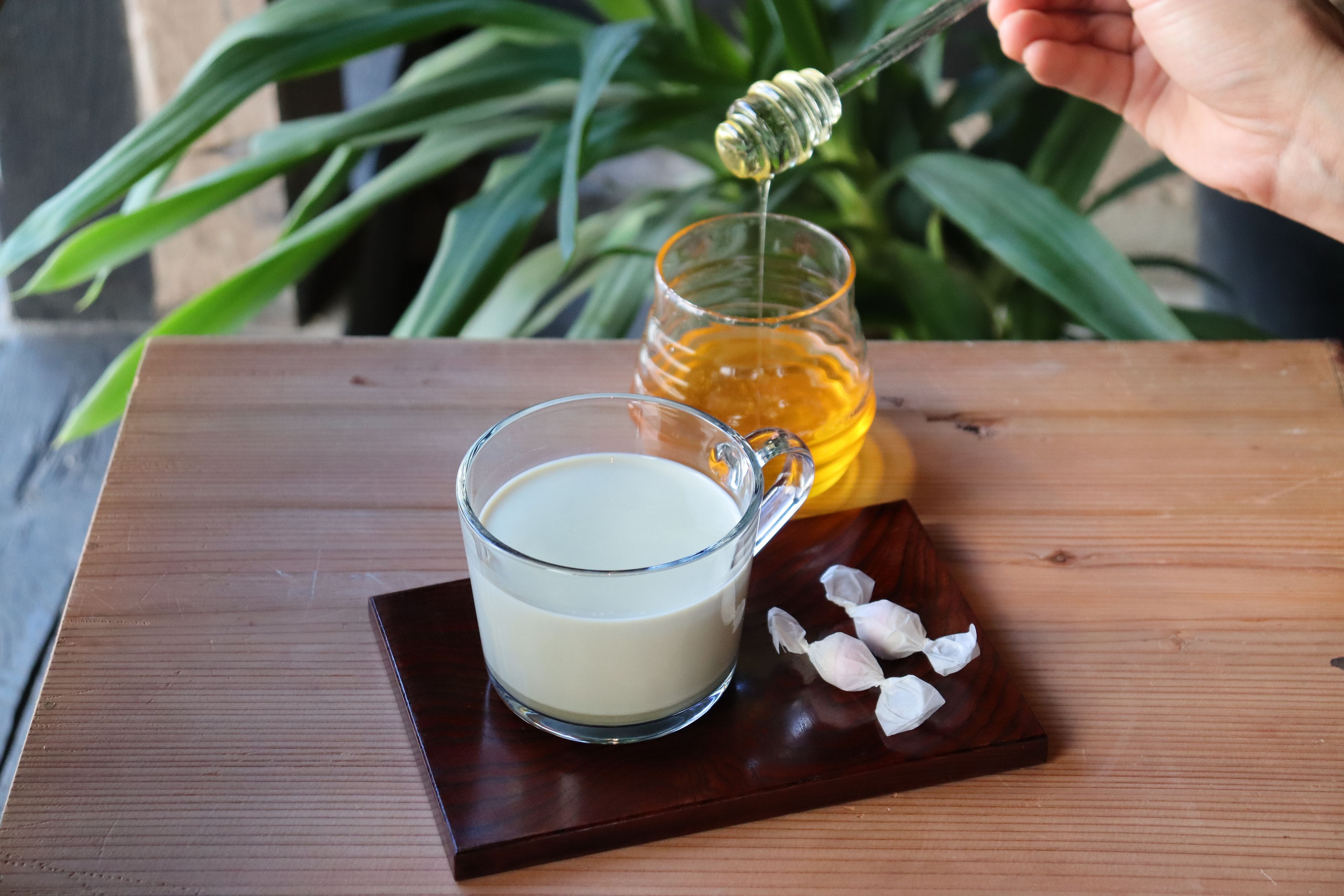 A wooden table with a glass cup of milk, a honey jar, and a honey dipper.