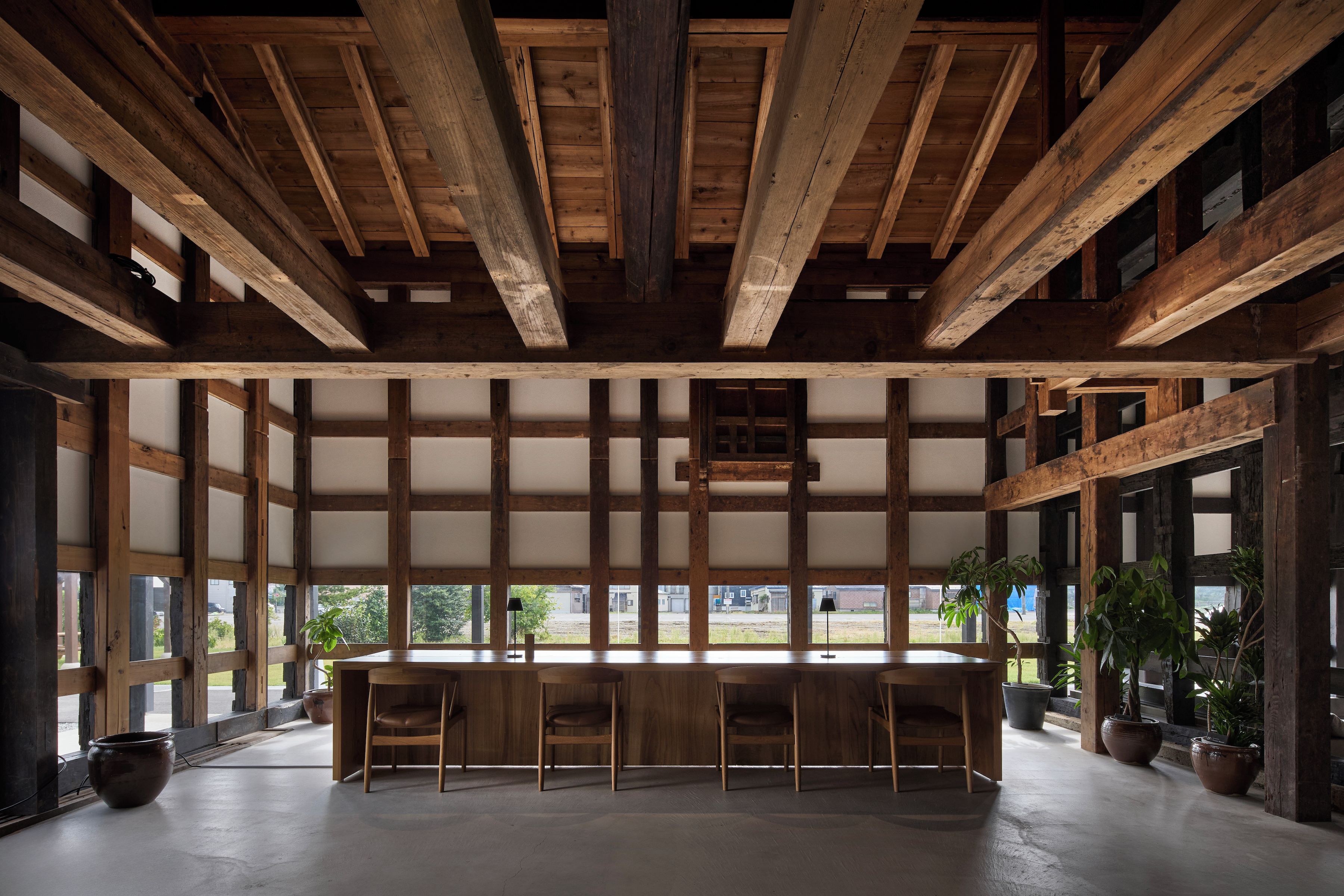 A spacious room with a high ceiling and exposed wooden beams. There is a long wooden table with chairs in the center, surrounded by large windows.