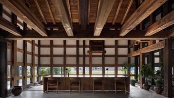 A spacious room with a high ceiling and exposed wooden beams. There is a long wooden table with chairs in the center, surrounded by large windows.