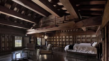 A spacious room with a large bed, two chairs, a glass table, and wooden beams on the ceiling.