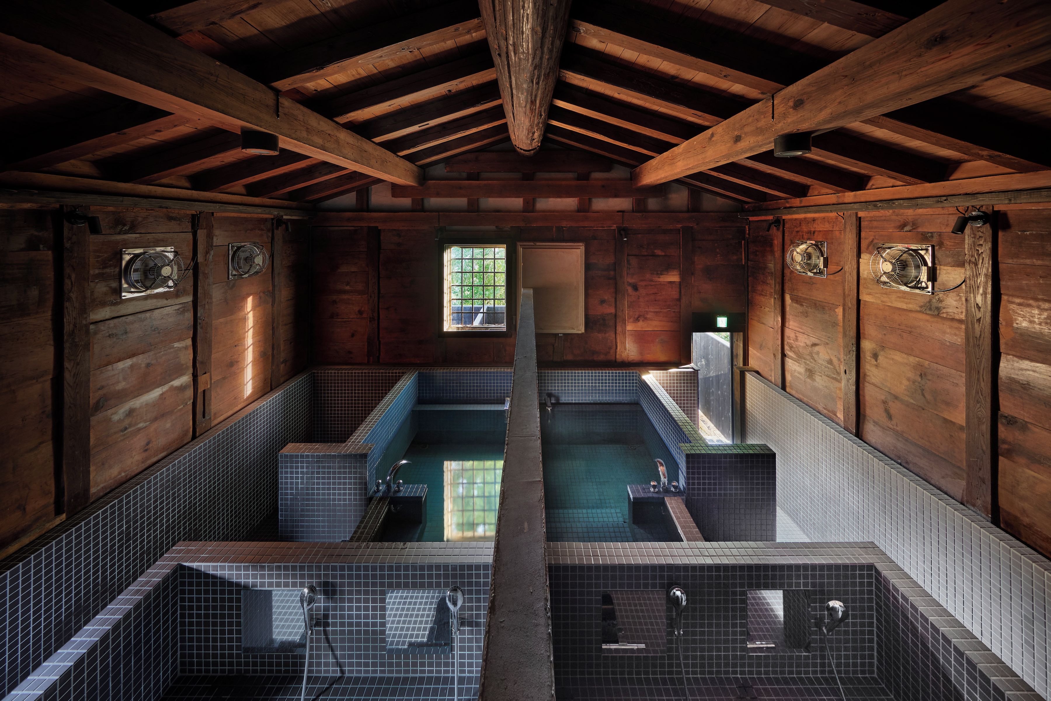 A wooden cabin with two separate bathing areas, each with a small window and a fan.