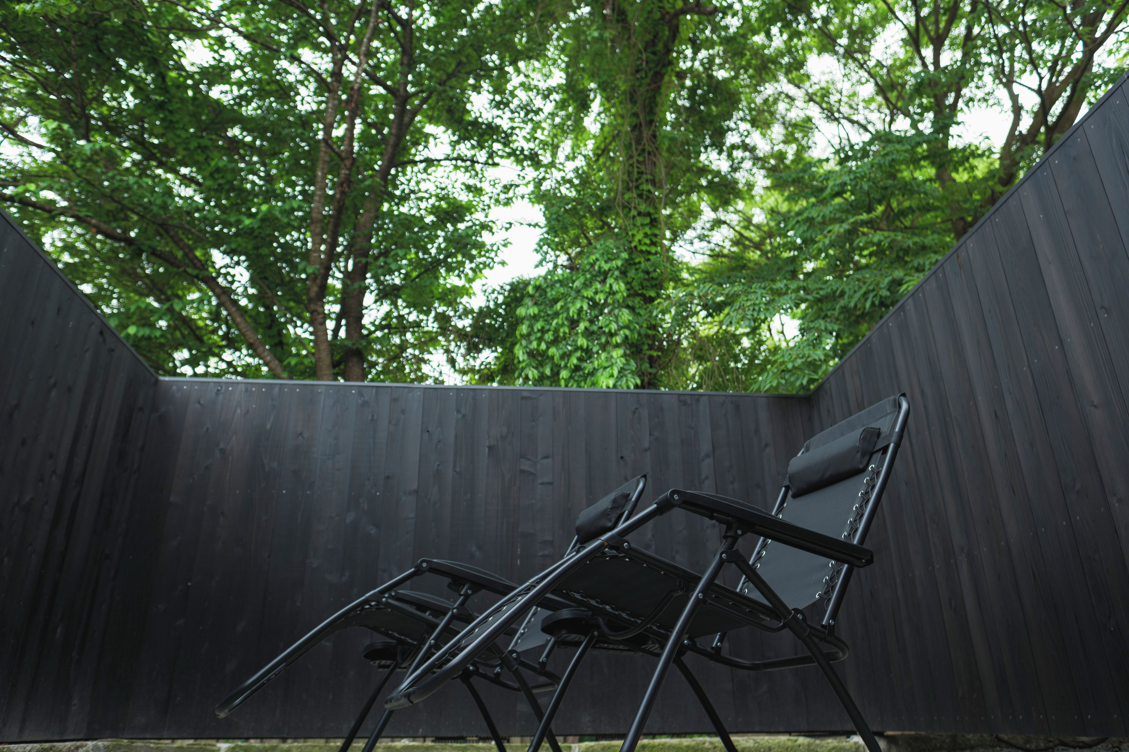 Una silla plegable negra en un espacio exterior con una cerca de madera y árboles al fondo.