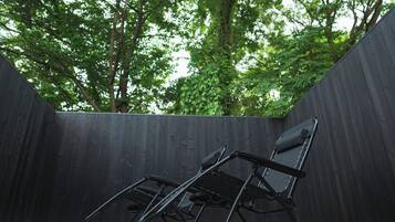Una silla plegable negra en un espacio exterior con una cerca de madera y árboles al fondo.