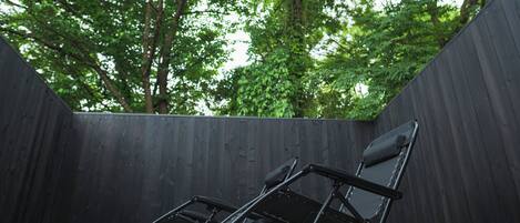 Una silla plegable negra en un espacio exterior con una cerca de madera y árboles al fondo.