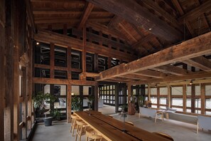 A spacious interior with high ceilings and exposed wooden beams, featuring a long wooden table and several chairs, surrounded by large windows and potted plants.