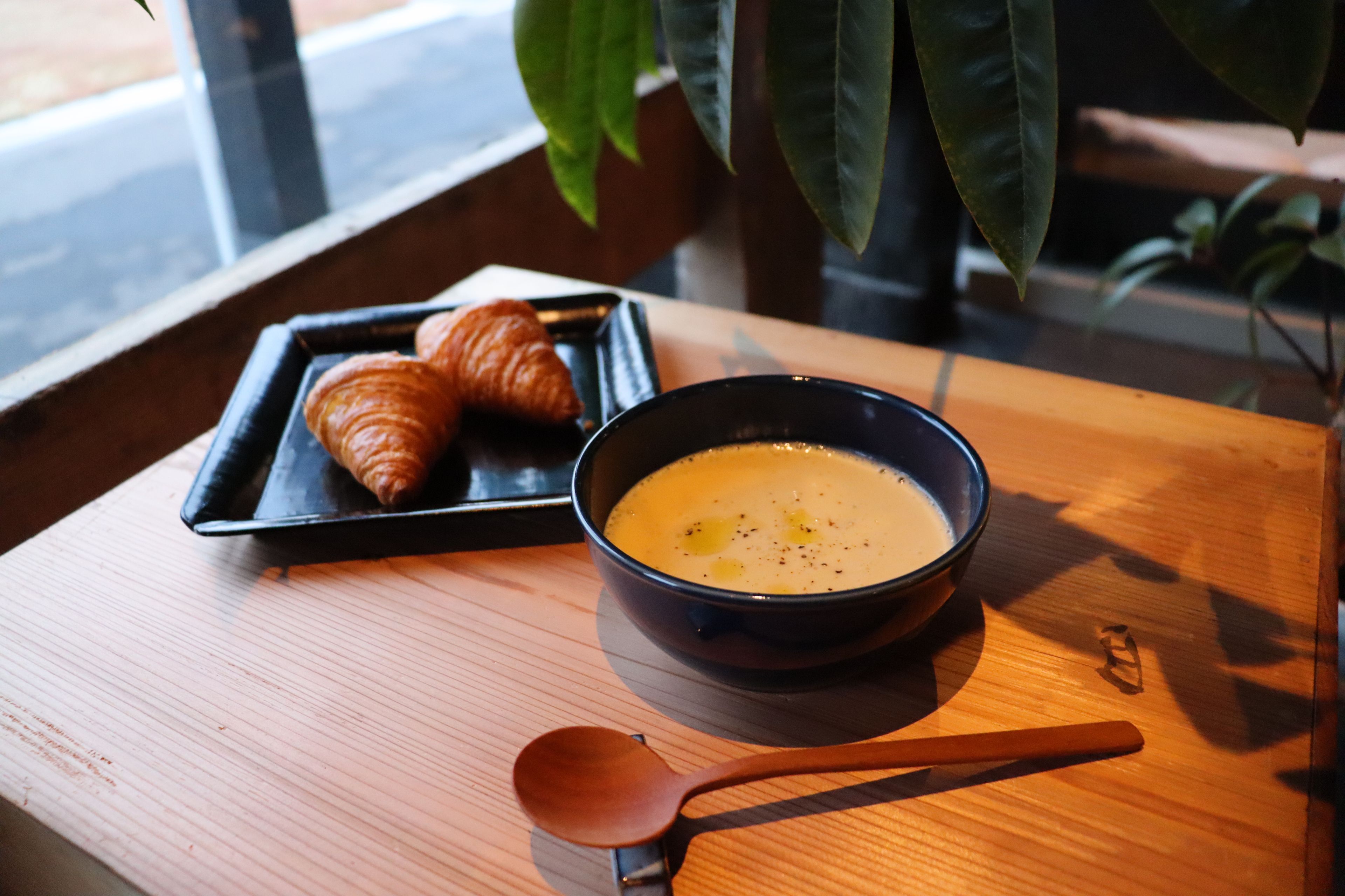 A tray with croissants and a bowl of soup on a wooden table near a window.