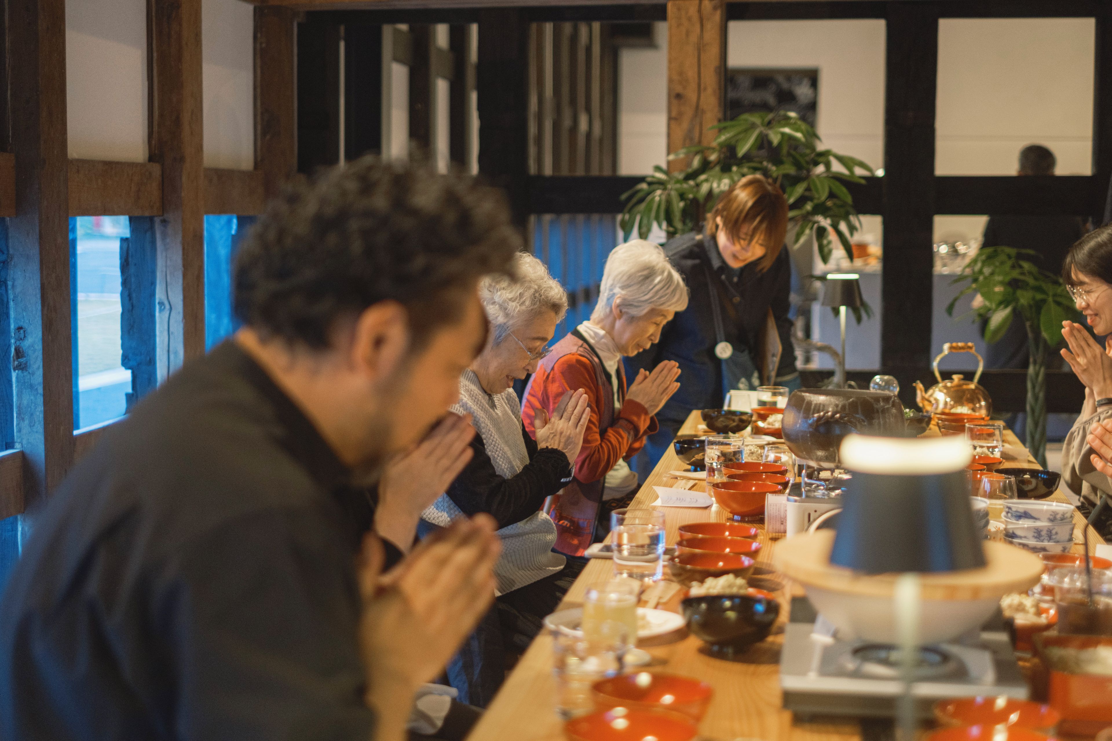 A group of people seated around a table with various dishes and a teapot, in a room with wooden beams and a plant.