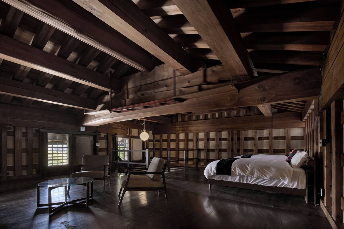 Una habitación amplia con una cama grande, dos sillas, una mesa de vidrio y vigas de madera en el techo.