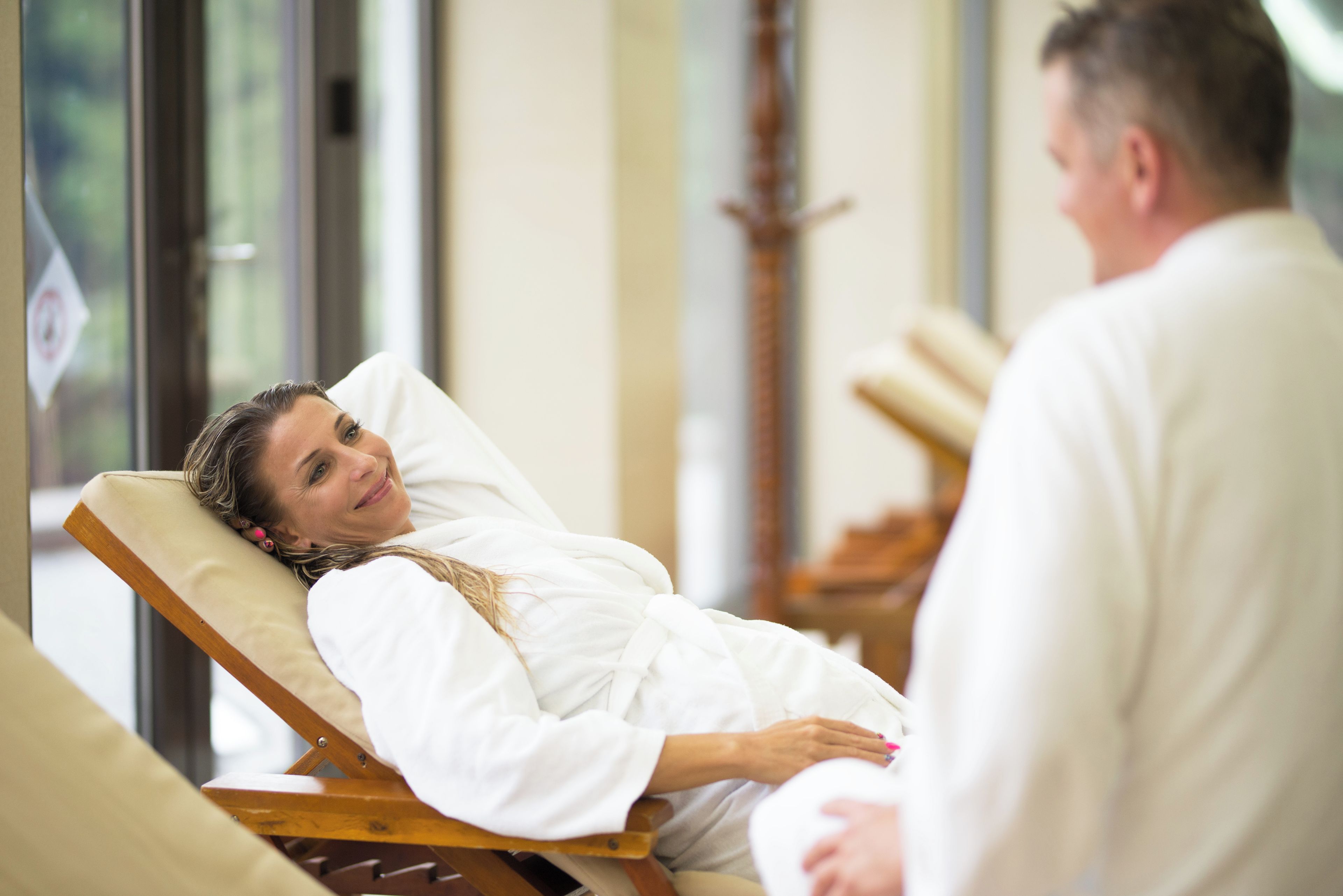 A person lying on a wooden chair in a room with a window, another person standing nearby, and a wooden structure in the background.