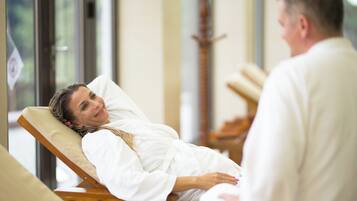 A person lying on a wooden chair in a room with a window, another person standing nearby, and a wooden structure in the background.