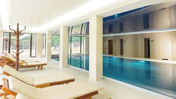 A modern indoor swimming pool with clear glass walls, sun loungers, and a wooden ceiling.