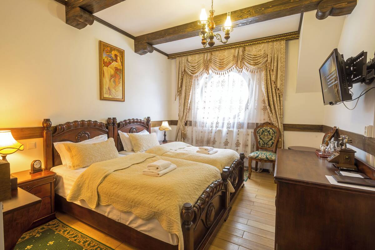A bedroom with a double bed, a wooden nightstand, a desk, a chair, a television, and a painting on the wall.