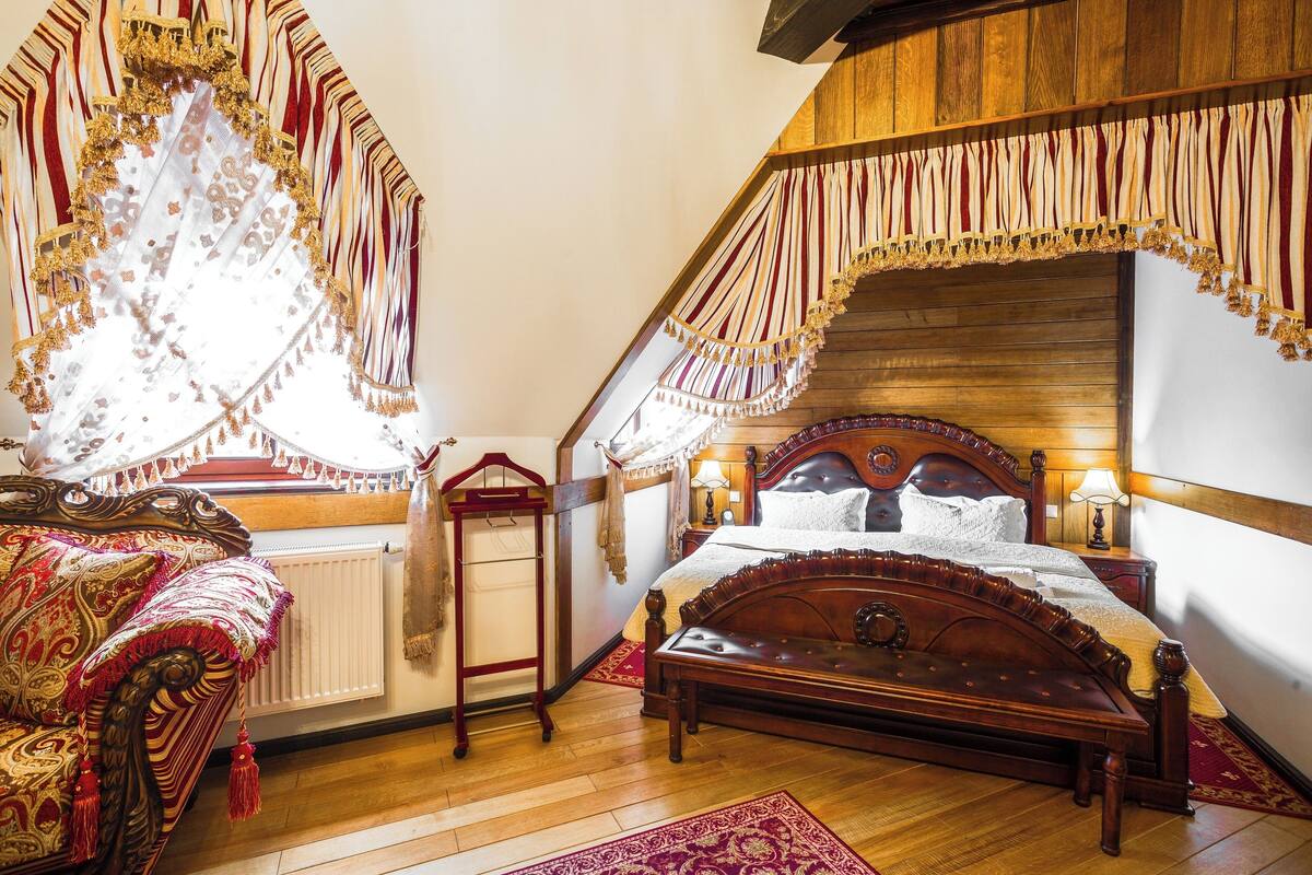 A bedroom with a wooden bed, a wooden bench, a radiator, and a window with curtains.