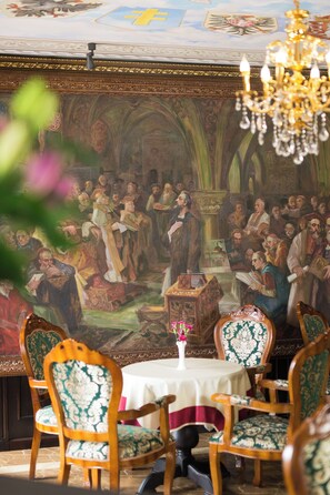 Un comedor con un mural, sillas ornamentadas, una araña y una mesa con un jarrón de flores.