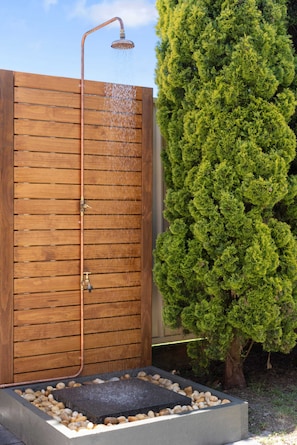 Eine Duschkabine aus Holz mit Regendusche, ein kleines Becken mit Kieselsteinen und ein hoher Nadelbaum.