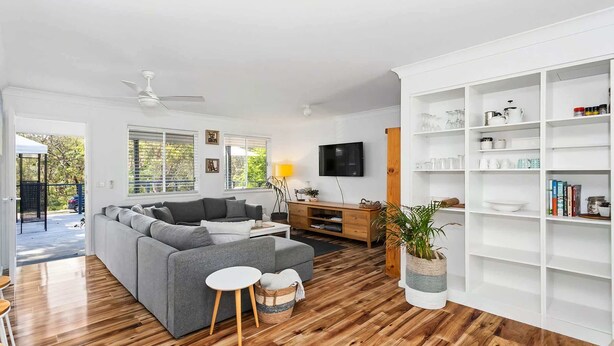 Ein modernes Wohnzimmer mit einem grauen Ecksofa, einem weißen Couchtisch und Holzboden. Es gibt eine weiße Regaleinheit mit diversen Gegenständen und einen an der Wand montierten Fernseher.