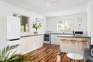 A modern kitchen with white cabinets, a built-in oven, a microwave, and a bar area with stools. - All Decked Out at Kioloa Pet Friendly (Kioloa)