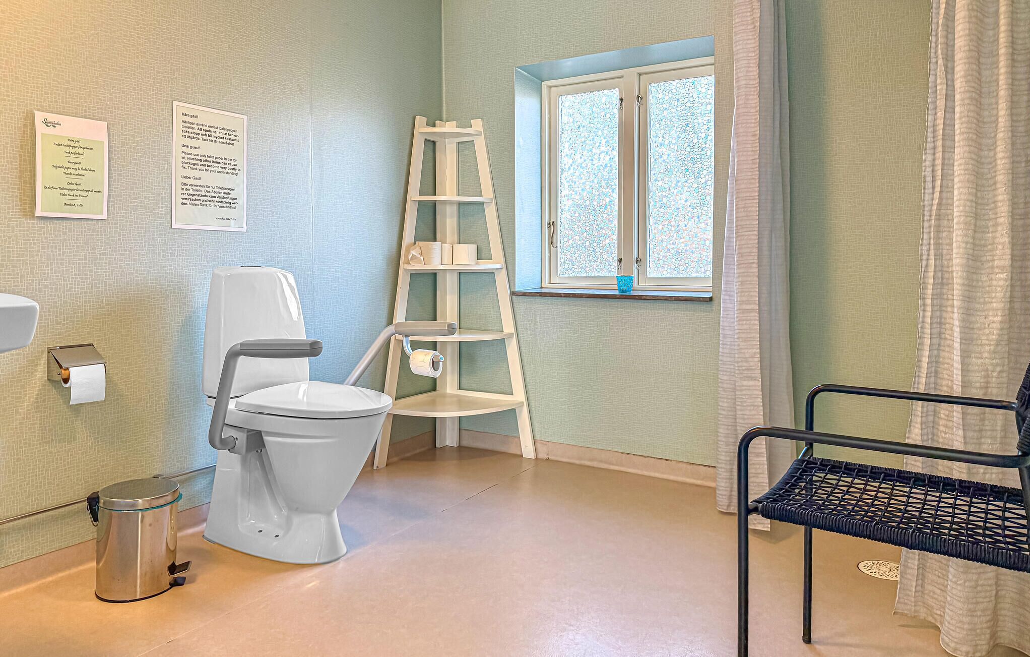 A bathroom with a toilet, a trash bin, a ladder, a window, and a chair.